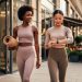 Two women in sustainable athleisure leave a studio onto a city sidewalk at golden hour, one carrying a yoga mat and the other adjusting a smartwatch, with a softly blurred cafe and greenery behind them